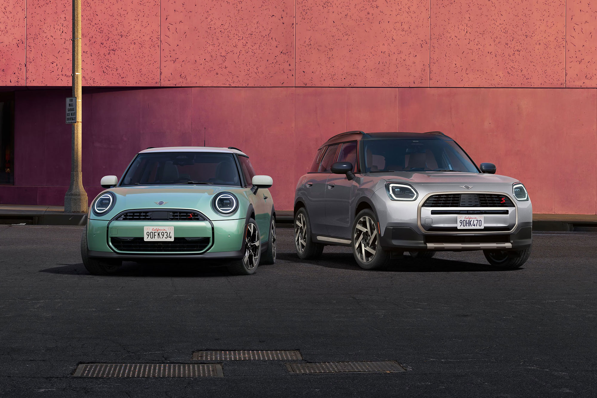 Front-view of the 2025 MINI Cooper S 2 Door in Ocean Wave Green and 2025 MINI Countryman S ALL4 in Melting Silver III parked side by side in front of a salmon-colored concrete building.